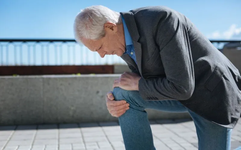 Elderly man bending forward while holding his knee in an outdoor setting, demonstrating an approach to prevent bone aches in cold weather through light exercises